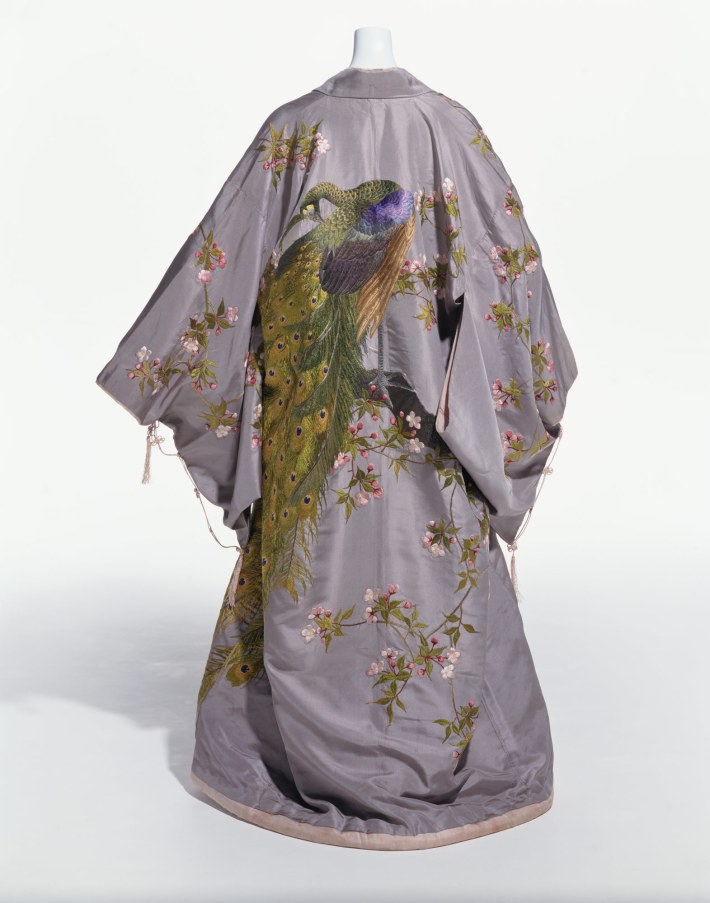 At-Home Gown (Kimono) by the Iida and Company, circa 1906. In the collection of, and image via,  The Kyoto Costume Institute. The green peafowl, or pavo muticus, is embroidered on this kimono by a leading Japanese export company. Originally from Burma (today’s Myanmar) in Central Asia, the green  peacock found its way to Japan in the seventeenth century and became embedded in the country’s artistic traditions. In  Japan, the peacock is a symbol of dignity and beauty; it is represented here alongside cherry blossoms, the eternal symbol for spring. In Europe and America, the Japanese kimono was  especially fashionable as an informal at-home garment like the tea gown or dressing gown.  